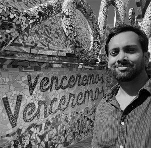 Black and white photo of Ashik Siddique, a young man with short black hair, a short beard, and brown skin. He is smiling in front of a wall with a elaborate mosaic reading Venceremos Venceremos.